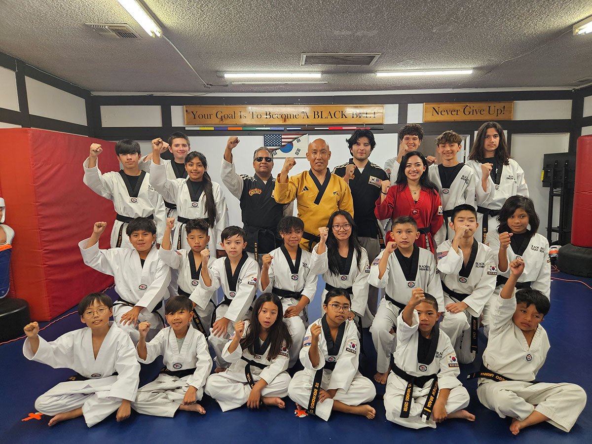 A group of Taekwondo students and instructors posing together with raised fists in front of dojo banners.