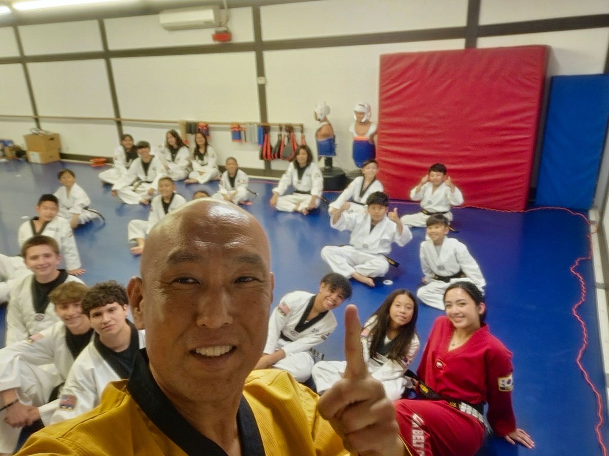 A smiling Taekwondo master in a gold uniform taking a high-angle selfie with a class of students sitting on the blue dojo mat.