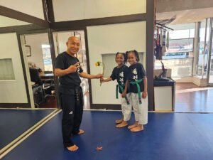A Taekwondo instructor in a black uniform smiling and making a peace sign while holding a sunflower with two young green belt students.
